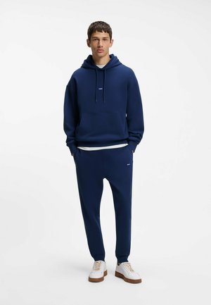 Ung mand iført en marineblå hættetrøje og matchende træningsbukser med hvide sneakers, stående mod en ensfarvet hvid baggrund.