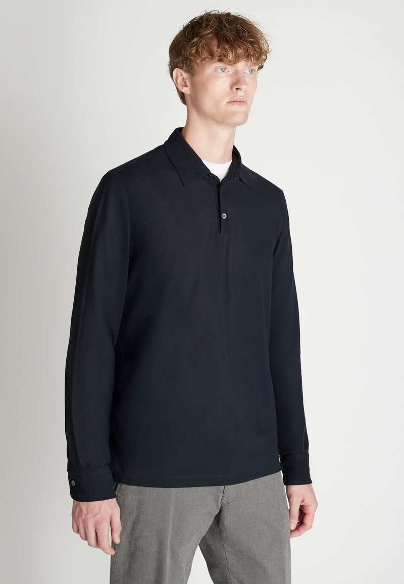 Schwarzes Langarm-Polohemd mit Kragen, Knopfleiste mit zwei Knöpfen und Bündchen. Glatte Textur, figurbetonte Passform, kombiniert mit grauen Hosen.