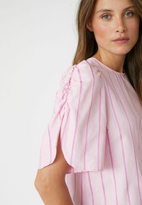Camicia rosa con strisce bianche verticali, dotata di maniche a sbuffo corte e scollo rotondo. Il tessuto ha una consistenza liscia.