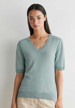 Ung kvinde med langt, glat hår iført en lysegrøn, kortærmet V-hals sweater og beige bukser, der står op ad en hvid væg.