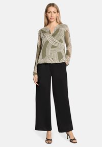 Durchscheinende olivgrüne Bluse mit weißem abstraktem Muster, V-Ausschnitt, lange Ärmel. Kombiniert mit schwarzen Hosen mit weitem Bein und schwarzen Riemchen-High Heels.