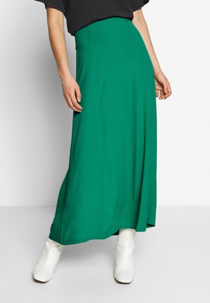 Femme portant une jupe verte taille haute, longue jusqu'au sol, avec des bottines blanches, debout devant un fond blanc uni.