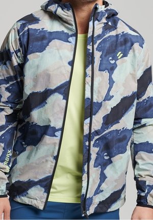 Homme portant une veste à capuche à motif camouflage bleu, gris et beige par-dessus une chemise vert clair, la veste étant partiellement zippée.