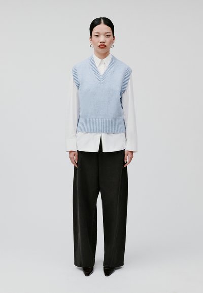 Lichtblauw gebreid vest met een V-hals, ribbels aan de randen, gedragen over een wit shirt. Gecombineerd met zwarte wijde broek en puntige zwarte schoenen.