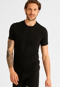 Svart T-skjorte med kort erme i bomull, med klassisk rund hals og tettsittende design, båret av en person med synlige tatoveringer på armen.