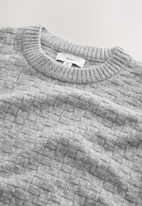 Maglione grigio lavorato a maglia con motivo a quadrati testurizzati, girocollo a coste e etichetta con dettagli del marchio e della taglia visibili.
