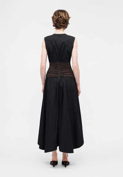 Ženska s kratkimi kodrastimi lasmi, oblečena v črno obleko brez rokavov z nagubanim rjavim pasom okoli pasu in črnimi čevlji z visokimi petami, stoji obrnjena stran.