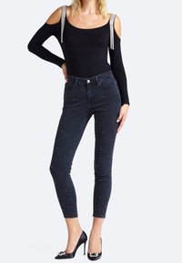Bodysuit negro con hombros al aire y tirantes plateados, combinado con jeans de mezclilla oscuros con adornos de perlas y un corte slim, llevado con tacones negros.