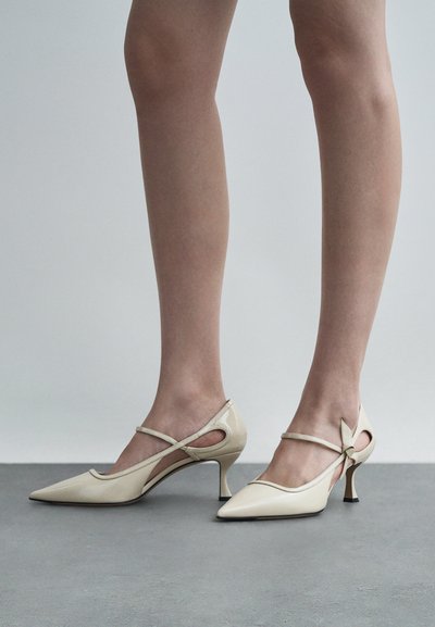 Μπεζ παπουτσοπέδιλα από πατέντα με ψηλό τακούνι και μυτερή μύτη, με κομμένα πλαϊνά και λεπτό λουράκι στον αστράγαλο. Έμφαση σε σκαλιστό τακούνι.