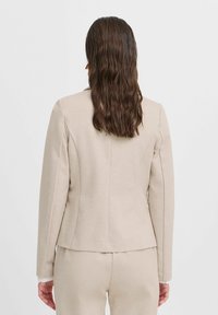 Krémově béžový přiléhavý blazer s dlouhými rukávy, elegantním designem, hladkou texturou a decentními detaily švů. Záda mají čistý finish.