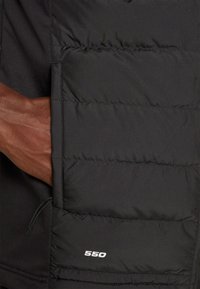 Main à peau foncée dans la poche d'une veste matelassée noire avec "550" cousu en blanc près du bord inférieur de la poche.