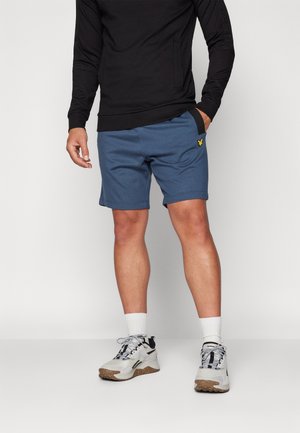 Mand iført blå shorts med gul logo, sort langærmet top, hvide sokker og hvide sneakers med sorte detaljer, der står mod hvid baggrund.