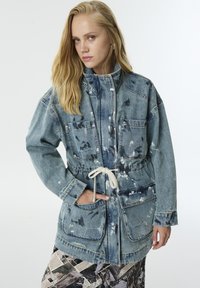 Blonde vrouw in een vervaagde blauwe denim jas met trekkoorden en verfspetters, met een patroon op haar broek, poserend met een hand in de zak.