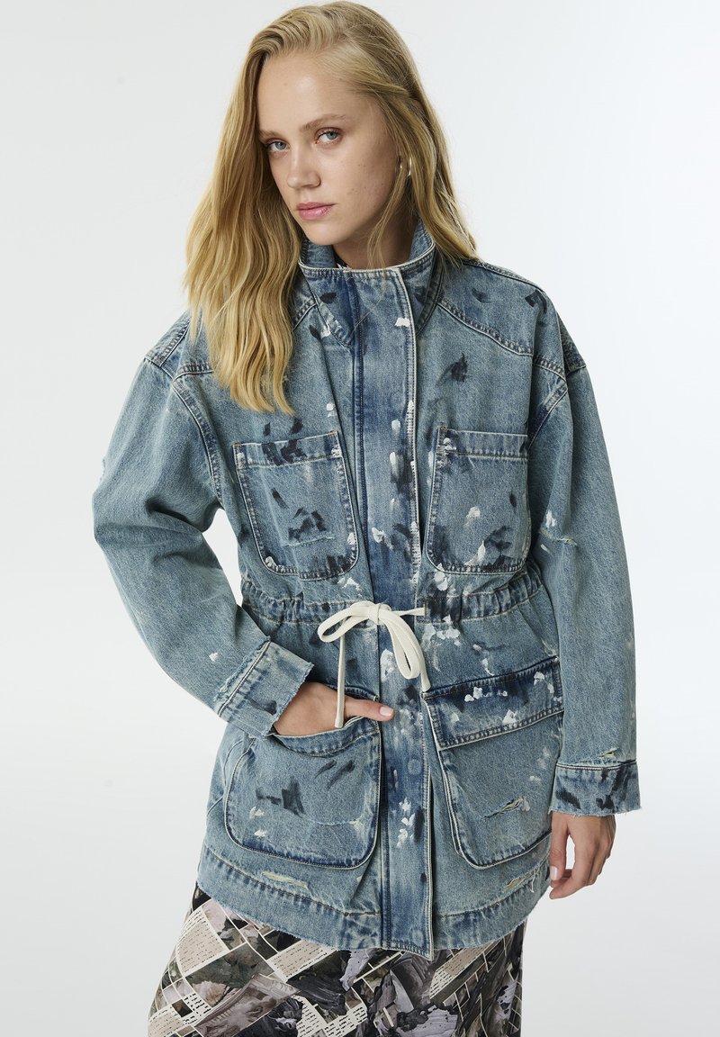 Blonde vrouw in een vervaagde blauwe denim jas met trekkoorden en verfspetters, met een patroon op haar broek, poserend met een hand in de zak.