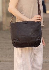 Dark brown leather shoulder bag with a soft texture, single adjustable strap, and a subtle logo on the front. Smooth, minimalist design.