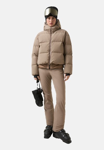 Beżowa pikowana kurtka z zamkiem błyskawicznym, czarne rękawice, jasnobrązowe spodnie narciarskie i czarne buty narciarskie; na głowie ciemna czapka z lustrzanymi goglami narciarskimi.