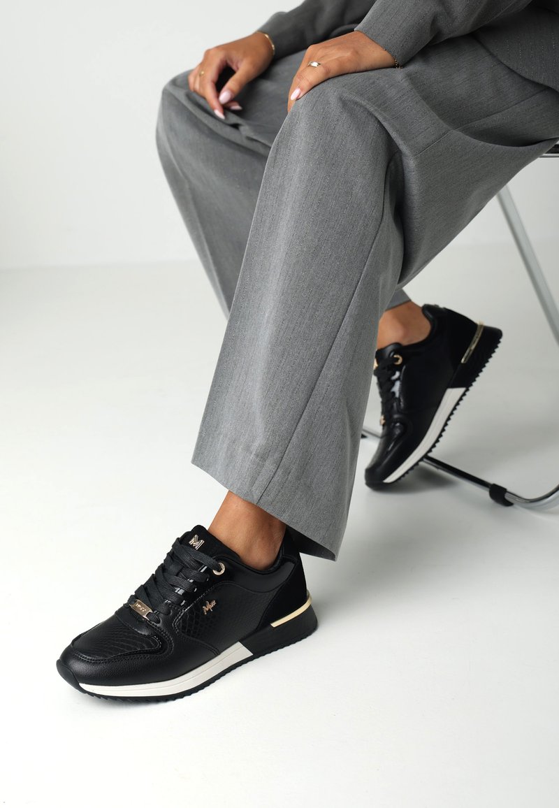 Baskets noires en cuir texturé, avec des accessoires en or et des accents blancs. Portées avec un pantalon gris sur mesure, montrant un design moderne et décontracté.