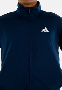 Marineblauwe ritsjack met hoge kraag. Voorzien van een wit logo met drie strepen op de linkerborst en een gladde stofstructuur.