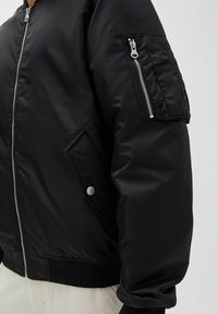 Veste bomber noire avec fermeture éclair, ourlet côtelé et poche latérale dotée de quincaillerie en argent et d'un détail à bouton-pression.