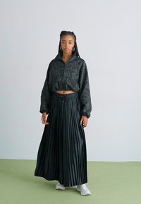 Sort cropped pullover med blomstermønster og høj krave, parret med en lang plisseret nederdel. Modellen har hvide sneakers på. Grøn gulv baggrund.