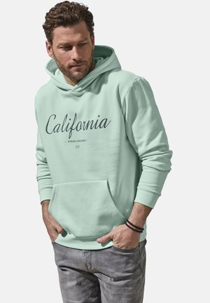 Mann trägt hellgrünen Hoodie mit dem Schriftzug „California State College“, graue Jeans und ein schwarzes Perlenarmband, Hände in den Taschen.