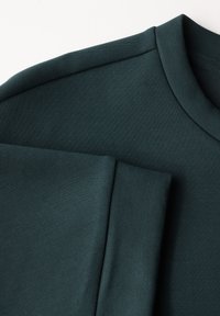 Tmavě modrozelené mikině s hladkou, texturovanou tkaninou. Design zahrnuje kulatý výstřih a detail skládání rukávů.