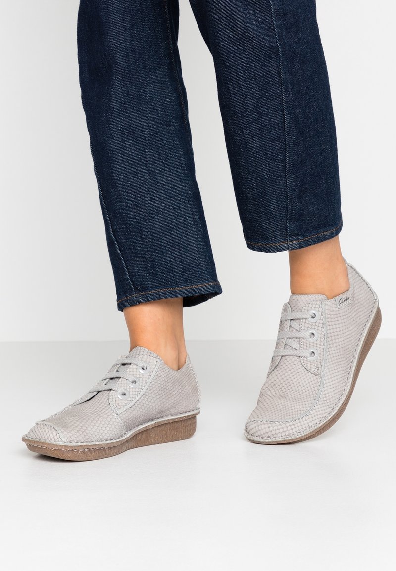 Personne portant des chaussures à lacets texturées grises avec des semelles marron et un jean bleu foncé raccourci, debout sur une surface blanche.