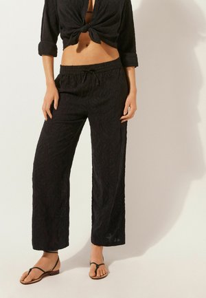 Blusa negra con estampado floral cruzada a la cintura, combinada con pantalones negros livianos y holgados. Sandalias negras minimalistas completan el conjunto.