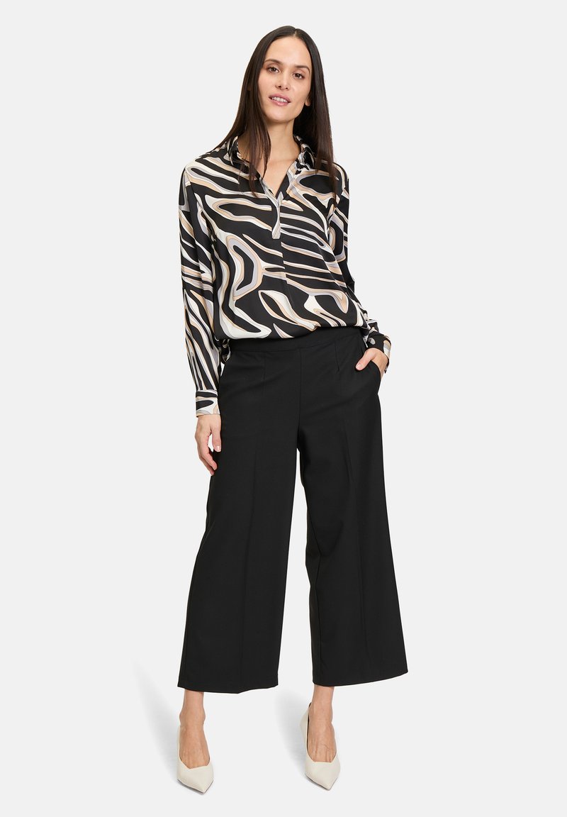 Blouse à motifs noir et crème avec un col, manches longues, associée à un pantalon large noir et des talons clairs.