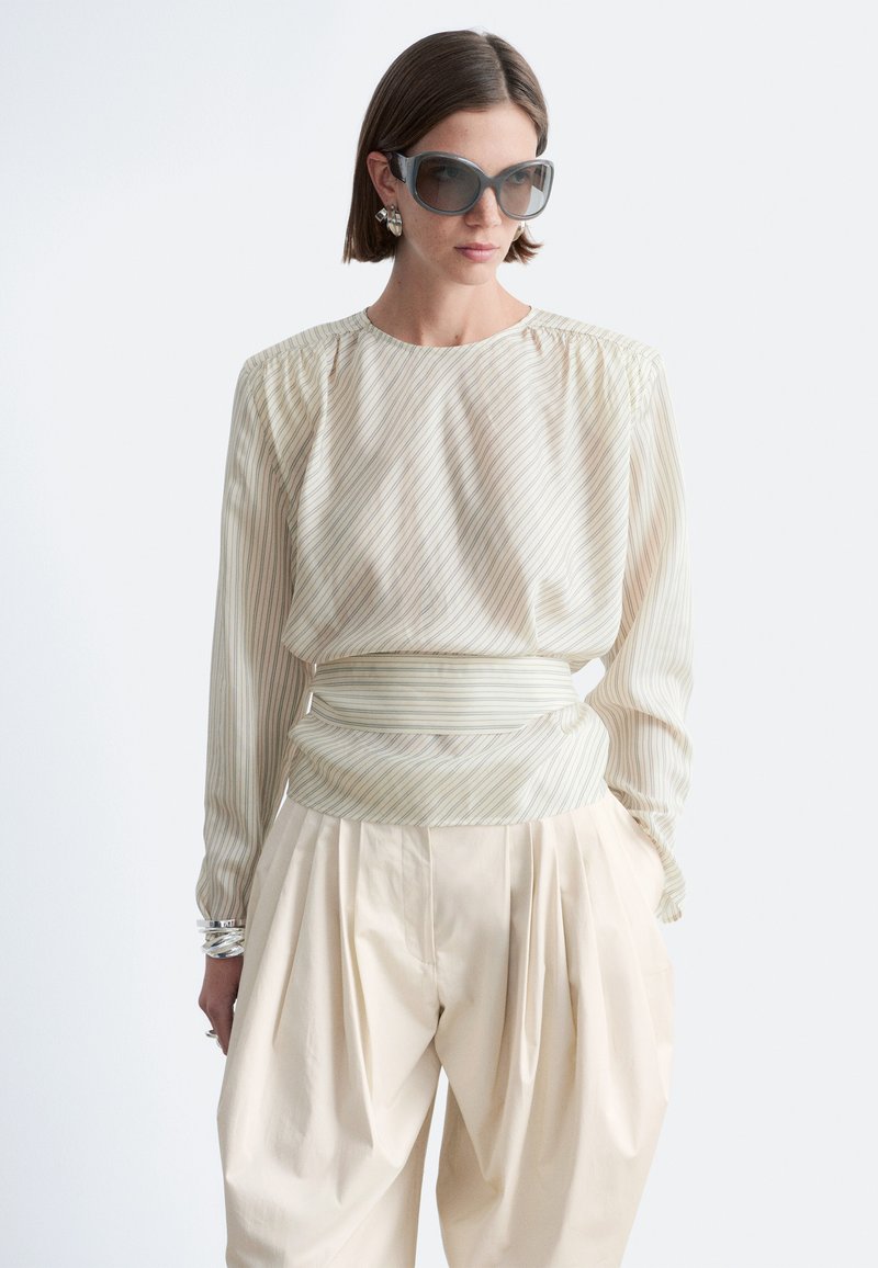 Femme aux cheveux bruns courts portant de grandes lunettes de soleil grises, un chemisier transparent rayé avec une large ceinture, un pantalon plissé crème et des bijoux argentés.