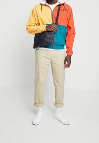 Giacca a vento color block gialla, arancione e teal. Chinos beige chiaro, sneakers bianche e una borsa a tracolla nera. Calzini bianchi visibili.