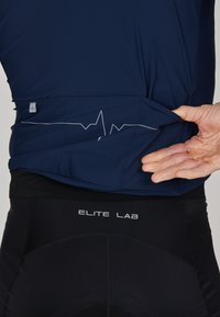 Marinefarbener Sportoberteil mit einer Herzschlaglinie auf dem Rücken; getragen über schwarzen Shorts mit dem "ELITE LAB"-Logo am Bund.