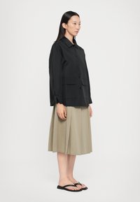 Camicia nera con bottoni e maniche lunghe, due tasche frontali, abbinata a una gonna plissettata color khaki e infradito neri, in piedi su uno sfondo neutro.