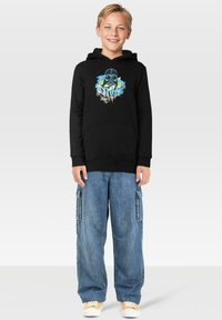 Sudadera negra con diseño gráfico que presenta una máscara y acentos coloridos, combinada con pantalones cargo de mezclilla azules holgados y zapatillas blancas.