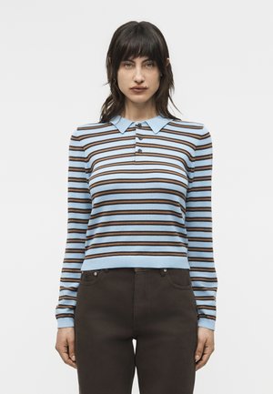 Nő világoskék és barna vízszintes csíkos hosszú ujjú ingben hegyes gallérral és sötétbarna nadrágban, karjai lazán állnak.