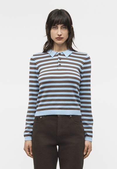 Femme portant une chemise à manches longues à rayures horizontales bleu clair et marron avec un col pointu et un pantalon marron foncé, debout les bras détendus.