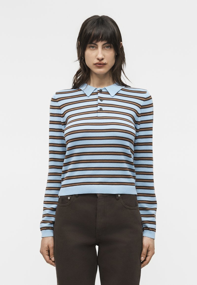 Femme portant une chemise à manches longues à rayures horizontales bleu clair et marron avec un col pointu et un pantalon marron foncé, debout les bras détendus.