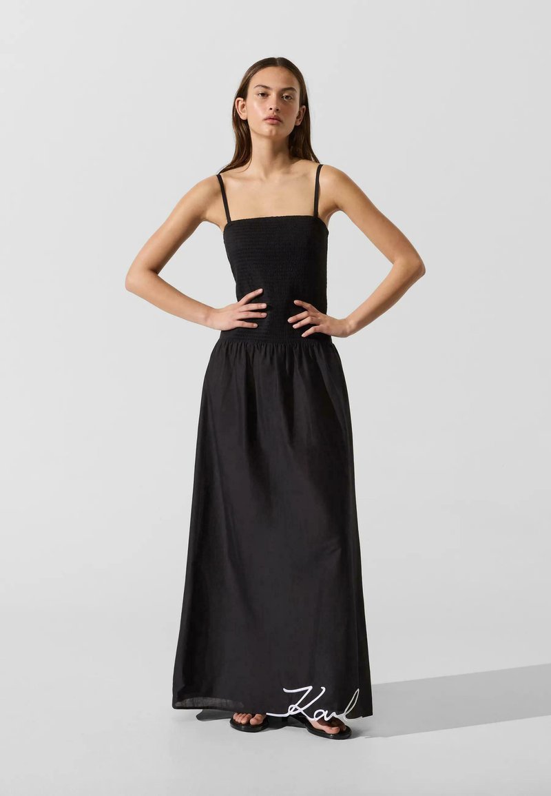Jeune femme debout, mains sur les hanches, portant une longue robe noire à fines bretelles et des sandales noires sur un fond gris uni.
