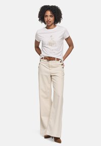 T-shirt blanc avec un motif abstrait doré, associé à un pantalon large crème avec des accents de boutons noirs et une ceinture marron.