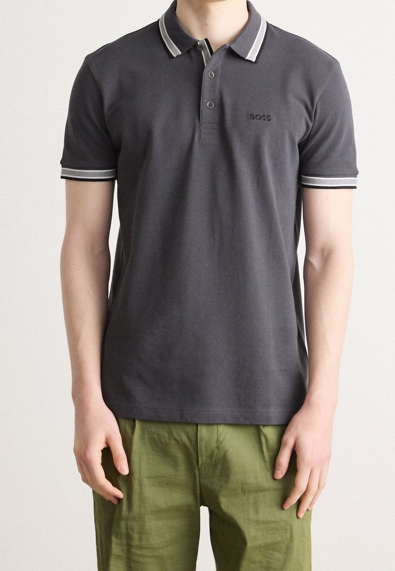 Homme portant un polo gris foncé avec col et manches rayés blancs, associé à un pantalon vert olive, debout devant un fond uni.