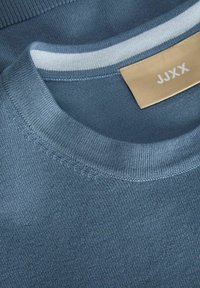 Maglione di cotone blu con collo rotondo, colletto a costine e un delicato motivo a righe. L'etichetta mostra il nome del marchio "JJXX" su uno sfondo beige.