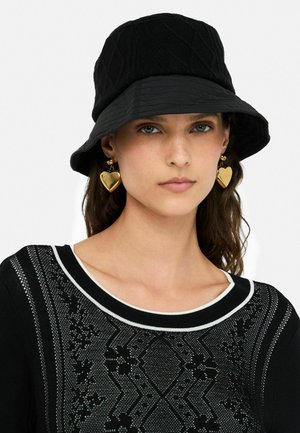 Cappello a secchiello nero con parte superiore in maglia texturizzata e visiera liscia. Indossato con orecchini a forma di cuore in oro e un maglione nero a fantasia.