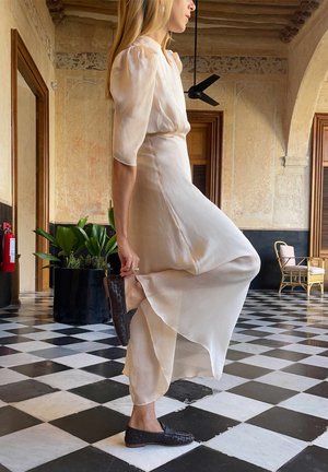 Woman in a light dress adjusting dark woven shoes on checkered floor in a historic building with arched doorways and potted plants.