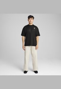 Sort kortærmet T-shirt med rund hals og logo patch på venstre bryst, parret med beige løstsiddende bukser og sorte sko.