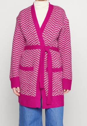 Femme portant un cardigan à motif chevron rose et beige avec une ceinture, sur un col roulé blanc et un jean bleu.