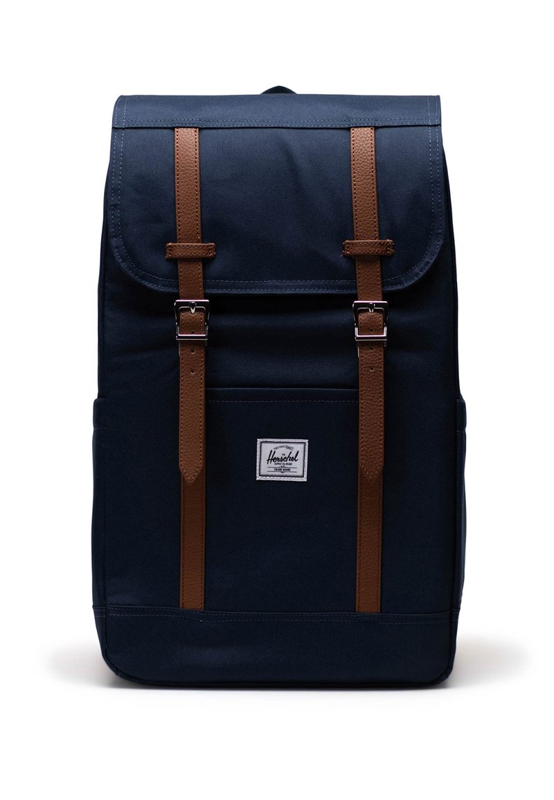 Marinblå canvasryggsäck med bruna läderremmar, utrustad med fälllock, justerbara remmar och en liten logopatch på framsidan.