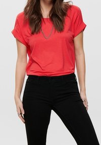 Korálové tričko s krátkým rukávem a ohrnutým lemem, měkká tkanina, uvolněný střih, kombinováno s černými džíny a stříbrným řetízkem.