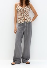 Canotta con motivo floreale in crema e rosa, con bordi arricciati e laccio frontale. Abbinata a jeans grigi a gamba larga e infradito nere.