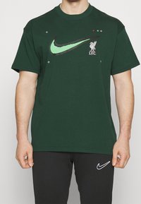 Zielony t-shirt z bawełny z dużym czarnym logo Nike oraz logo Liverpool FC, z kolorowymi akcentami i krótkimi rękawami.