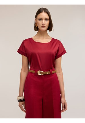 Vrouw met steil bruin haar draagt een rode satijnen top met korte mouwen, bijpassende broek, een zandkleurige riem en twee armbanden aan haar linker pols.